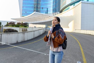 Şehir caddesinde sırt çantalı sıradan bir gezgin kadın, atkı ve ceket, modern mimari zemin, sokak çizgileri ve kentsel seyahat atmosferini ön plana çıkarıyor.