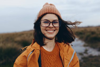 Gözlüklü, örgü şapkalı ve turuncu kazaklı mutlu bir kadın açık havada rüzgarlı havadan ve bulanık doğal manzaradan hoşlanıyor. Yaşam tarzı, sonbahar kıyafeti ve neşeli bir ruh hali..