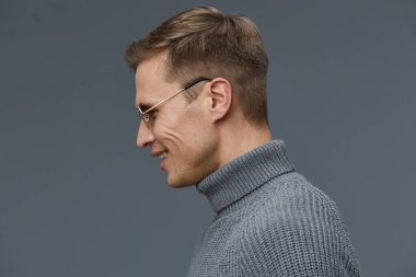 Gözlüklü ve gri balıkçı yaka kazaklı, gri arka planda duran gülümseyen beyaz adamın profili. Rahatlamış ruh hali zihinsel sağlık bilincini ve alet kullanımını ifade eder.