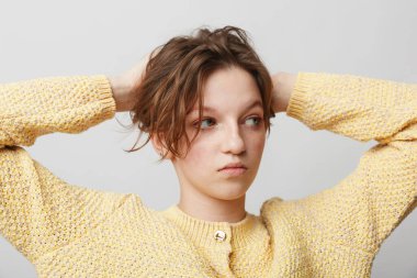 Sarı örgü süveter giyen, elleri başının arkasında olan genç bir kadının portresi. Sakin bir ifadeyle, sıradan moda ve hafif ışıklandırmayla yan tarafa bakıyor..