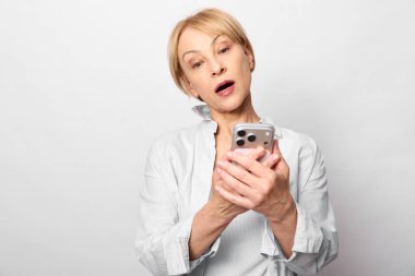 Beyaz bluzlu, kısa saçlı olgun bir kadın akıllı telefonu şaşırmış gibi tutar. Beyaz arka plan stüdyosu. Dijital iletişim, duygular ve modern teknoloji kullanımı kavramı.