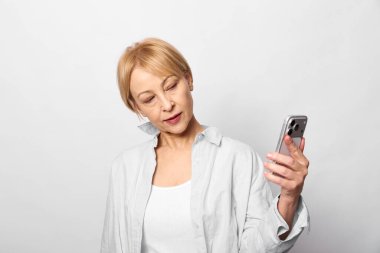 Orta yaşlı, kısa sarı saçlı bir kadın akıllı telefonu tutuyor ve ekrana ilgiyle bakıyor. Gündelik beyaz gömlek ve nötr arkaplan modern teknoloji kullanımını vurguluyor.