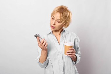 Kısa saçlı, açık renk tişörtlü, elinde kahve fincanı olan ve akıllı telefon ekranını kontrol eden bir kadın. Beyaz arka planda izole edilmiş. Modern teknoloji kavramı, rutin ve gündelik tarz..