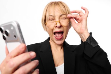 Kısa sarı saçlı ve gözlüklü mutlu orta yaşlı kadın akıllı telefon tutuyor ve beyaz arka planda açık ağızlı bir ifadeyle neşeli bir selfie çekiyor. Dijital bağlantı ve eğlence kavramı.