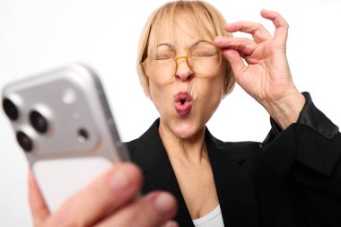Gözlüklü ve siyah ceketli gülümseyen orta yaşlı bir kadın akıllı telefonlu selfie çeker, komik bir yüz ifadesi takınır ve göz kırpar. Beyaz arkaplan, günlük tarz, sosyal medya ve kendini ifade kavramı.