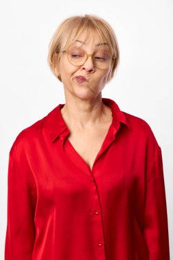 Kafası karışmış, gözlüklü, kırmızı bluzlu orta yaşlı bir kadın beyaza izole edilmiş şüpheli bir yüz ifadesi gösteriyor. Tereddüt, şüphe, sorgulama ve belirsiz duygular kavramı.