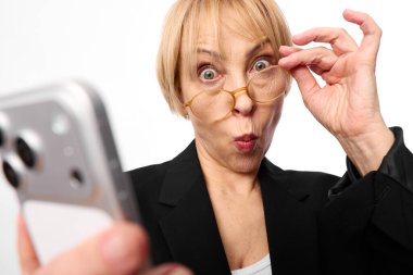 Siyah ceketli sarışın kadın şaşırmış yüz ifadesi takınırken ve beyaz arka planda selfie için akıllı telefon tutarken gözlüklerini ayarlıyor. Teknoloji, iletişim, selfie ve dijital kültür kavramı.
