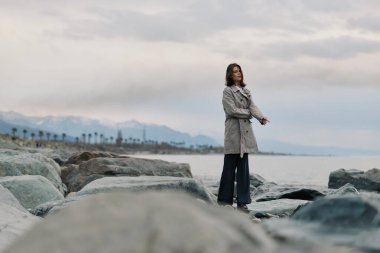 Paltolu ve siyah pantolonlu genç bir kadın deniz kenarındaki büyük kayalarda duruyor. Dağ manzaralı bulutlu hava ve arka planda palmiye ağaçları barışçıl bir atmosferi çağrıştırıyor.