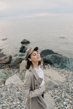 Woman in trench coat and cap stands on rocky shore with eyes closed, feeling calm and serene near vast water. Concept of nature, relaxation, and peaceful outdoor moment.