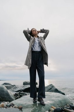 Palto ve pantolon giyen şık bir kadın elleri başının arkasında Rocky Beach 'te poz veriyor. Bulutlu gökyüzü ve okyanus arka planı gevşek ve zarif dış mekan stilini vurguladı.