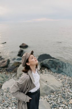 Woman wearing hat and loose cardigan breathes deeply on pebble shore near calm sea. Natural environment and casual fashion create mood of calmness and outdoor leisure.