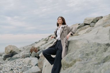 Uzun palto ve siyah pantolon giyen şık genç bir kadın bulutlu gökyüzünün altında sakin denizin yanındaki kayalık sahilde poz veriyor. Sıradan şehir tarzı ve doğal taştan arka planı olan sakin bir açık hava ortamı.