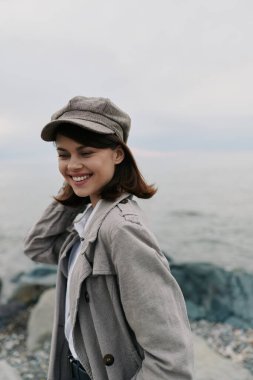 Happy young woman wearing grey coat and newsboy cap stands by rocky beach with ocean in background. Casual style and natural light create a relaxed, joyful outdoor atmosphere.