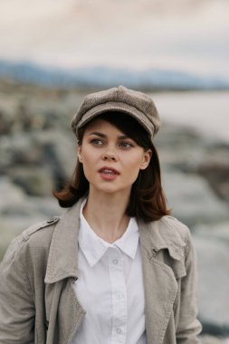 Kaliteli bir şapka takmış genç bir kadının portresi ve kayalık sahilde arkası bulanık su ve dağlarla kaplı hafif bir ceket. Açık hava yaşam tarzı ve mevsimsel stil kavramı.