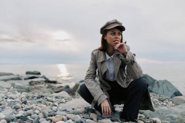 Paltolu ve şapkalı şık bir genç kadın bulutlu gökyüzü ile deniz kenarında çakıl taşlarının üzerinde oturuyor. Sonbahar stili, doğa ve açık hava ortamının düşünceli havası..