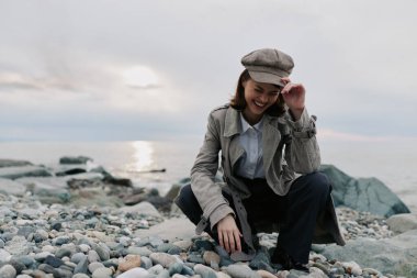 Retro ceket ve şapka giymiş mutlu genç bir kadın sakin sularda çakıl taşı kumsalında çömeliyor. Bulutlu gökyüzü ve doğal ışık açık hava yaşam tarzı için barışçıl bir atmosfer yaratır..