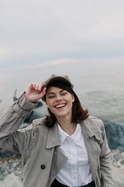 Smiling young woman in checkered coat and hat poses outdoors near rocky shore. Calm ocean and cloudy sky enhance serene mood. Concept of lifestyle, joy, and autumn fashion portrait.