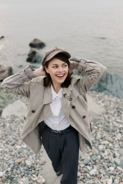 Smiling woman with coat and hat poses on pebble beach by calm sea. Casual autumn fashion, relaxed mood, natural environment and outdoor lifestyle captured in bright daylight near water.