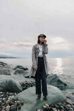 Uzun paltolu, şapkalı ve koyu renk pantolonlu zarif genç kadın arka planda bulutlu gökyüzü ve sakin deniz kıyılarında poz veriyor. Moda, doğa ve kıyı yaşam tarzı kavramı.