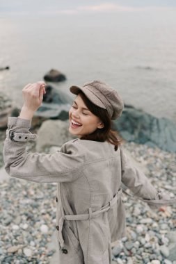 Hafif trençkotlu ve şapkalı gülümseyen kadın Rocky Sahili yakınlarında rüzgarlı bir günün tadını çıkarıyor. Mutluluk kavramı, gelişigüzel stil ve sakin sularda doğal soluk renkler..