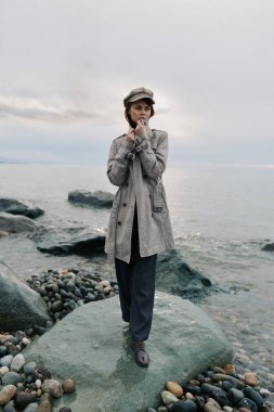Gri paltolu ve şapkalı genç kadın bulutlu gökyüzünün altında sakin deniz kenarında kayalık sahilde poz veriyor. Doğal ışık, sonbahar havası ve huzurlu sahil atmosferli açık hava moda portresi..