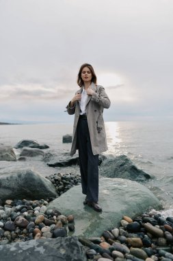 Kayalık sahilde, bulutlu gökyüzünün altında sakin okyanus kenarında şık kıyafetler giyen bir kadın. Doğal manzaralı kasvetli bir atmosfer. Sonbahar modası, açık hava yaşam tarzı ve kıyı çevresi kavramı.