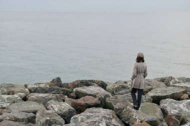 Ceket giyen yalnız bir kadın deniz kıyısında büyük kayalıklarda yürür. Bulutlu gökyüzü ve sakin su, doğal ortamdaki sessiz yalnızlığı ve iç gözlem havasını vurgular..
