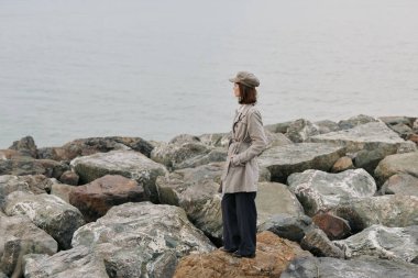 Bej ceketli ve şapkalı bir kadının sakin deniz kıyısındaki büyük kayalıklarda dikilirkenki görüntüsü. Sessiz atmosfer, gündelik moda ve bulutlu gökyüzü yansıma ve doğa bağlantısını çağrıştırıyor..
