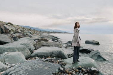 A thoughtful woman wearing a trench coat stands on rocky shore near the sea. Pebble stones and cloudy sky set a peaceful atmosphere in this natural coastal landscape scene.
