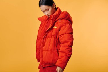 Parlak turuncu kışlık ceketli kadın model, sarı arka planda başı öne eğik poz veriyor. Eşofman, kaban, soğuk iklim, sıcak kıyafetler ve sokak modası konsepti için günlük kıyafet..
