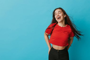 Kırmızı gömlekli ve siyah pantolonlu neşeli bir kadın mavi arka planda gözleri kapalı gülümsüyor. Genç, neşeli, günlük kıyafet ve doğal saç hareketi pozitif duygu ve özgürlüğü gösteriyor..