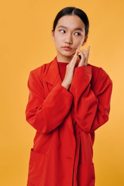 Asian female model dressed in oversized red jacket posing with contemplative look and hand near cheek on vibrant yellow backdrop for fashion or lifestyle concept.