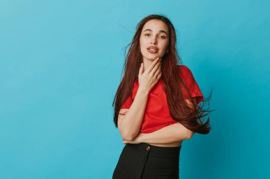 Kırmızı bluz ve siyah pantolon giyen genç bir kadının portresi mavi arka planda duruyor. Uzun saçlı, rahat giyimli, kendine güvenen, doğal ışık ve basit stüdyo ortamlı bir manken..