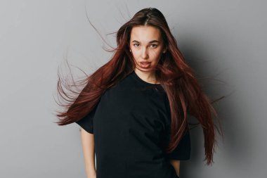 Siyah tişörtlü, dalgalı saçlı genç kadın yoğun bir görünüş ve doğal makyajla arka planda yumuşak ışıklandırma ve dinamik saç hareketleriyle..