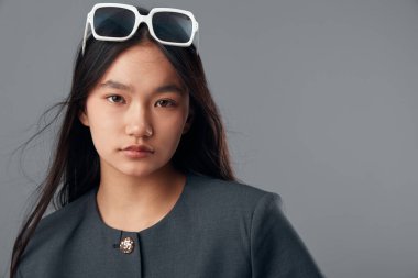 Doğal makyajlı, düz saçlı, beyaz gözlüklü ve resmi koyu ceketli güzel bir Asyalı kadın model. Gri duvardaki stüdyo resmi için poz veriyor..