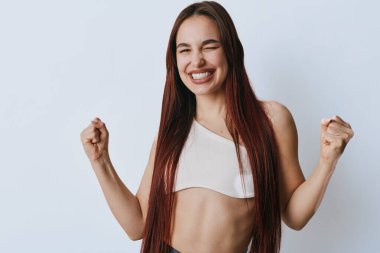 Uzun saçlı ve kaslı genç bir kadın. Yumruklarıyla sevinçten havalara uçuyor. Kadın, güler yüzlü ve enerjik. Sağlık ve kendine güven için hafif arka planda..