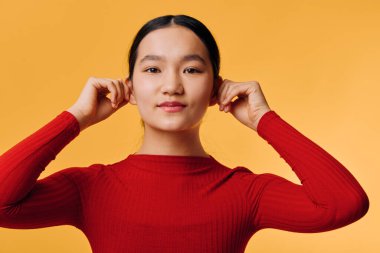 Female model in red long sleeve top fixing hair, posing with relaxed face and direct gaze on solid orange backdrop, closeup studio shot with soft lighting.