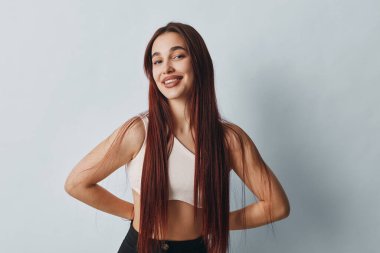 Smiling young female with long straight hair standing with hands on waist. Wearing sleeveless top and black pants, posing confidently in bright minimalist studio setting.