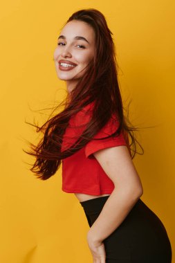 Smiling female model with flowing hair in red shirt and black pants posing sideways on yellow backdrop. Youthful woman showing positive emotion with natural look and relaxed posture.