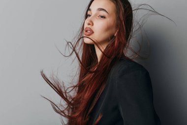 Siyah ceketli ve tarafsız makyajlı kızıl saçlı genç bir kadının portresi. Moda fotoğrafçılığında gri zemin üzerinde saç hareketi ve yumuşak ifade.