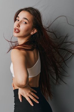 Beyaz kolsuz üst ve siyah alt kısımları olan uzun saçlı genç kadın modelin portresi. Gri arkaplanlı stüdyo çekimi doğal güzelliği ve rahat duruşu gösteriyor..