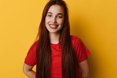 Smiling young woman with long brown hair standing in front of yellow backdrop wearing casual red t shirt. Positive female portrait with hands on hips and friendly face in bright setting.