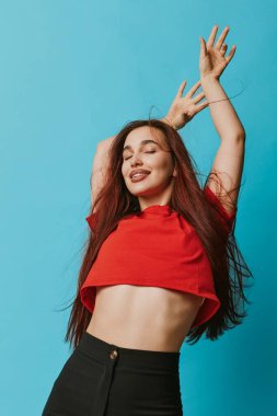Kırmızı bluzlu, siyah pantolonlu, kolları açık, gözleri kapalı genç bir kadın. Doğal gülüşlü ve rahat pozlu bir portre, yaşam tarzı ve moda için sade mavi bir zemin üzerinde..