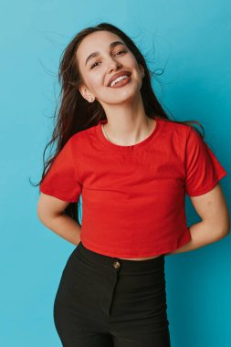Kırmızı bluzlu ve siyah pantolonlu gülümseyen kadın elleri arkada mavi arka planda duruyor. Uzun saçlı genç bir kadın günlük kıyafet portresine duyduğu güveni ve güveni ifade ediyor..