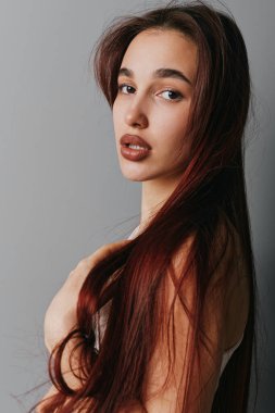 Young woman posing with long straight hair and neutral expression on gray backdrop. Closeup beauty shot with natural look, clear skin, and soft light for portrait or cosmetic concept.