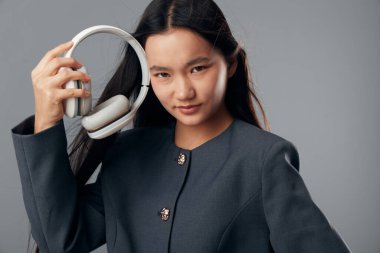 Uzun saçlı, kendine güvenen genç bir bayan stüdyoda kablosuz kulaklık tutuyor. Dinleme ya da dijital ses için koyu renk ceketli ve teknolojik aksesuarlı modern günlük kıyafet..