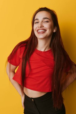 Happy young female with long straight hair in red crop top and black pants posing with eyes closed and wide smile on yellow studio background with confident posture.