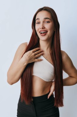 Uzun saçlı, sıradan giyimli, eli göğsünde ve belinde duran mutlu genç bir kadının portresi. Doğal makyajlı çekici bir kadın ve beyaz arka planda rahat bir ruh hali..