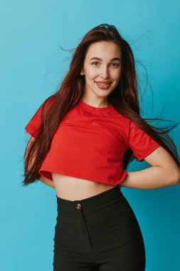 Smiling female model with long dark hair dressed in red crop top and black high waist pants standing with hands on hips on blue backdrop. Youthful style and happy expression in photo.