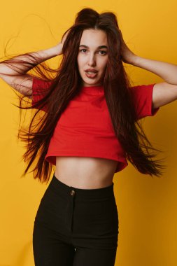 Portrait of confident girl in red shirt and black pants with flowing hair posing with hands on head on bright yellow backdrop for style and youth.